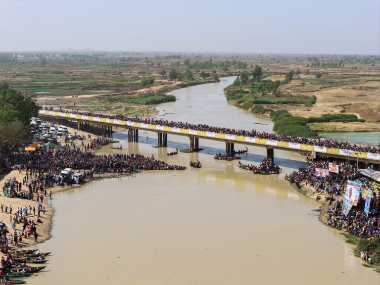 Argungu Fishing Festival Aerial view of the Matan Fada River during Argungu’s youth water sports day, with audience on the riverbank and the Argungu–Birnin Kebbi bridge in the background.