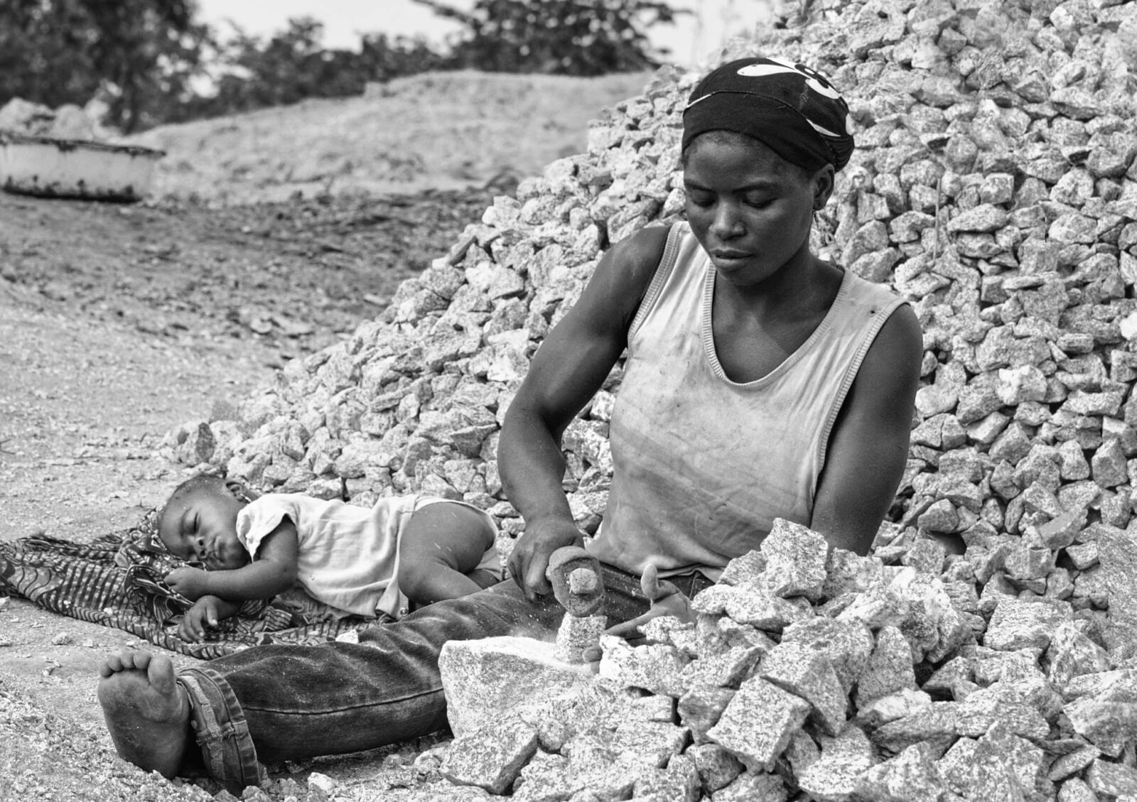 A mother breaks stones for construction in Gudi, Nasarawa State, Nigeria, while her baby sleeps beside her amid dust and heat.