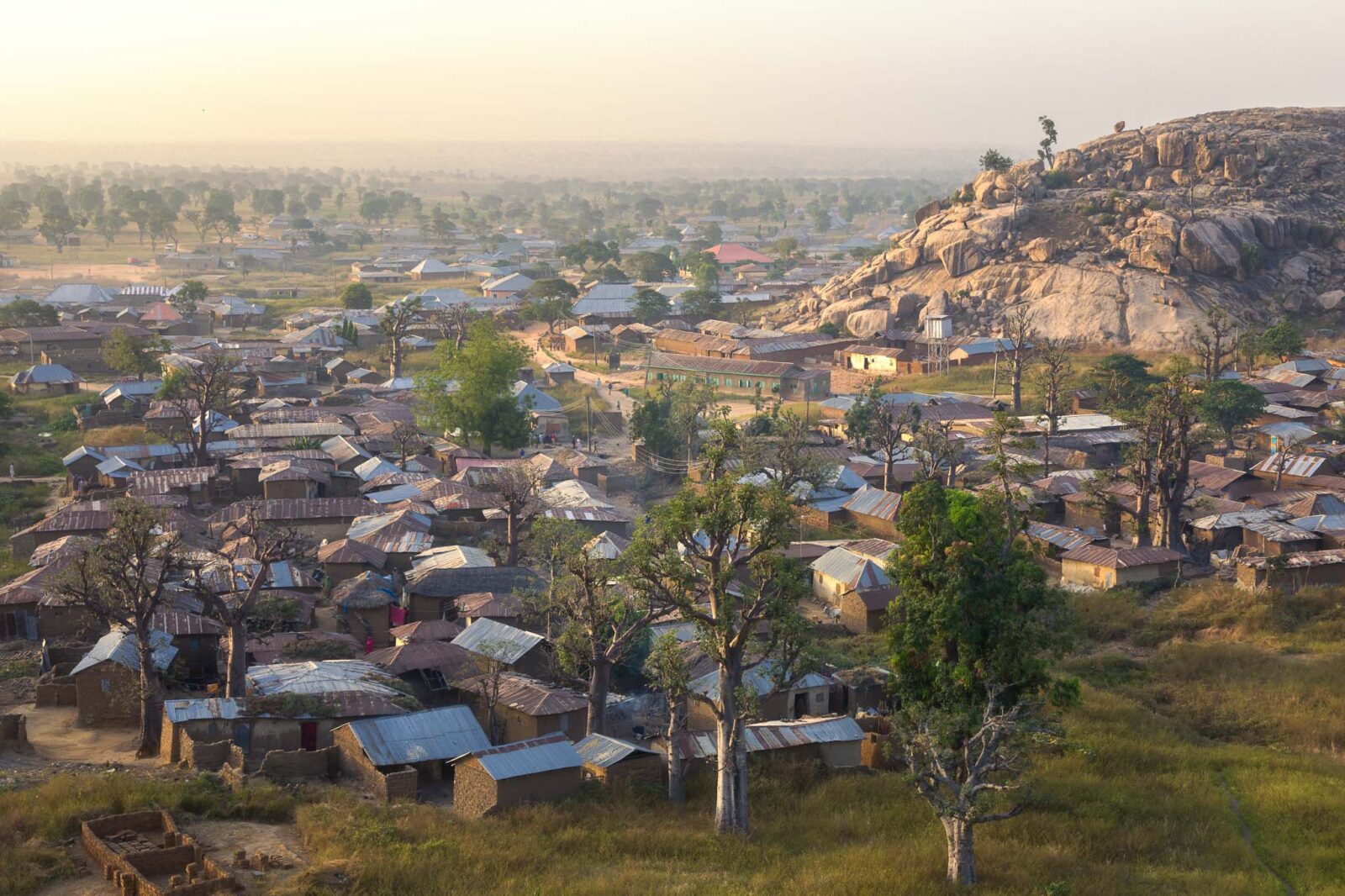 Turunku village seen from a hill at dawn, with mud houses, tin roofs, baobab trees, foggy savannah, and a rocky mountain.