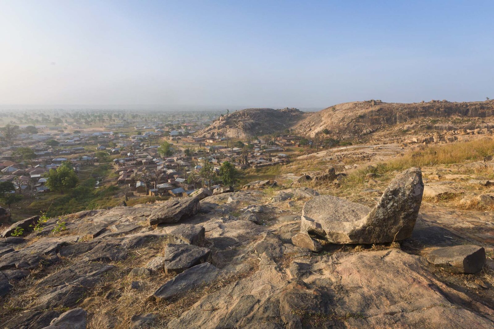 Wide-angle view of Turunku village from hilltop with Queen Amina’s Stone Seat in the foreground, mud houses, baobab trees, foggy savannah, and rocky mountain.