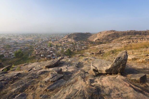 Queen Amina’s Stone Seat Overlooking Turunku Village – Wide-Angle Dawn View | Irene Becker Photography | Irene Becker Photography Wide-angle view of Turunku village from hilltop with Queen Amina’s Stone Seat in the foreground, mud houses, baobab trees, foggy savannah, and rocky mountain.