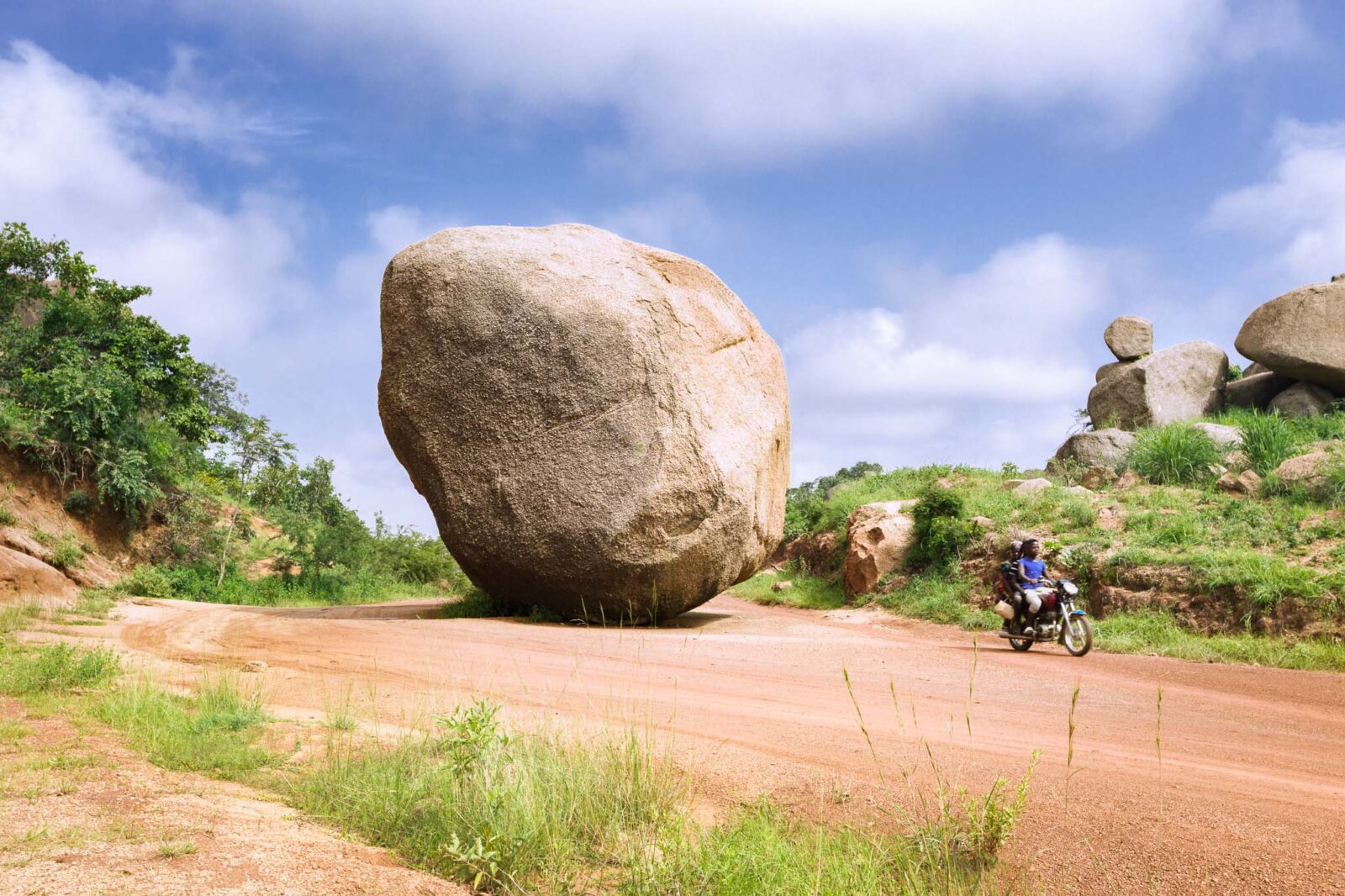 A motorcyclist rides from Gengere village on a road in Maraban Kajuru, Kaduna State, Nigeria. A huge round rock divides the road, apparently fallen from the adjacent mountain.